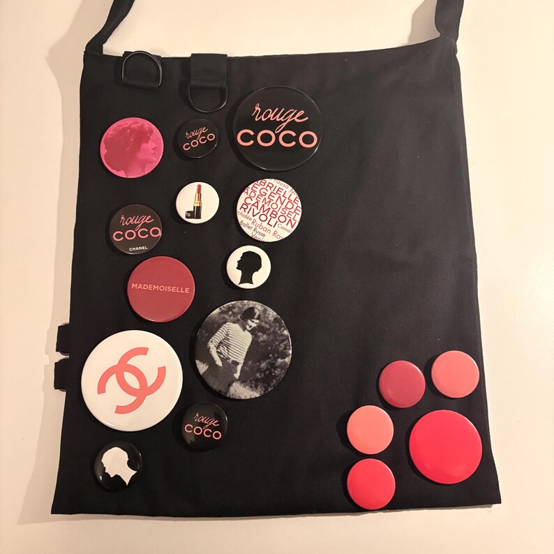 May include: Black tote bag adorned with various round pins. The pins feature text like "rouge coco" and "MADEMOISELLE", along with a Chanel logo and other graphics. The bag has a shoulder strap and a simple design.