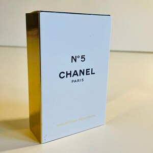 May include: A rectangular white box with gold-colored sides. The front of the box features the text "N&deg;5 CHANEL PARIS" in black. The bottom of the box has the text "COLLECTION S&Eacute;DUCTION" in gold.