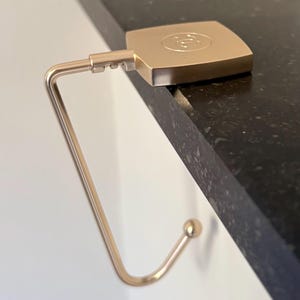 Può includere: Un gancio per borsa color oro con una parte superiore quadrata e un gancio curvo. La parte superiore ha un design circolare al centro. Il gancio è progettato per essere appeso al bordo di un tavolo.