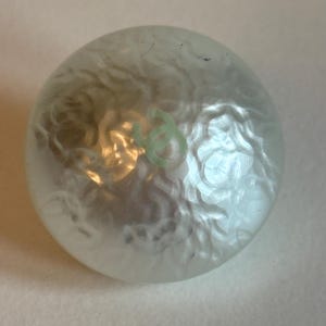 May include: A round, pearlescent button with a swirling, textured surface. A small, light green symbol is visible on the button's surface. The button has a slightly domed shape and reflects light, creating highlights and shadows.