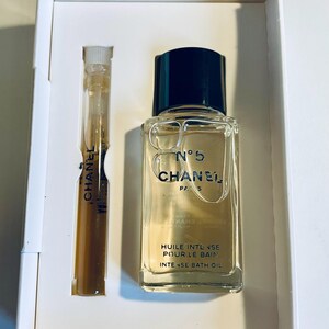 May include: A clear glass bottle of Chanel No. 5 Intense Bath Oil with a black cap, and a small vial of perfume. The bottle has the text "N&deg;5 CHANEL PARIS" and "HUILE INTENSE POUR LE BAIN" printed on it. The vial has "CHANEL" printed on it.