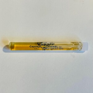 May include: A clear glass vial containing a golden-colored liquid. The vial has black text that reads "CHANEL PARIS" and "CHANEL INC. NEW YORK, N.Y. 10019". The vial is lying horizontally on a white surface.