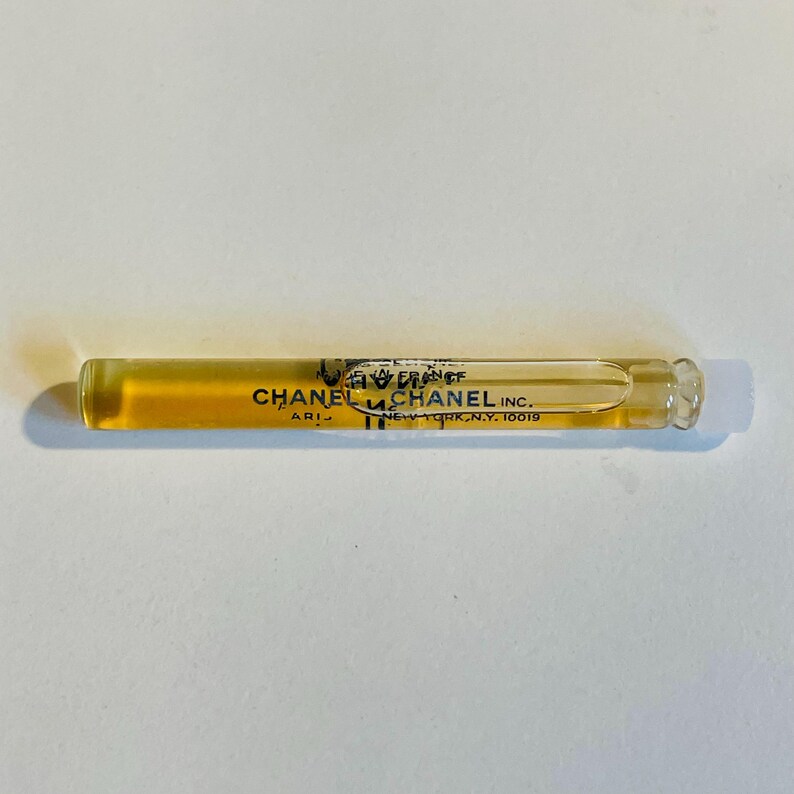 May include: A clear glass vial containing a golden-colored liquid. The vial has black text that reads "CHANEL PARIS" and "CHANEL INC. NEW YORK, N.Y. 10019". The vial is lying horizontally on a white surface.