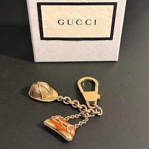 May include: A gold-toned Gucci keychain with a horse racing helmet and a small orange purse charm. The keychain is attached to a gold clasp and chain. The Gucci logo is visible on the clasp and the box in the background.