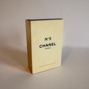 May include: A rectangular, cream-colored box with the text "N&deg;5 CHANEL PARIS" in black. The box also has the words "COLLECTION SEDUCTION" printed on it. The box is standing upright on a white surface.