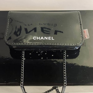 Puede incluir: Un monedero negro brillante con una cadena plateada. La palabra "CHANEL" est&aacute; impresa en blanco en la parte delantera. El monedero es rectangular y est&aacute; sobre una superficie negra reflectante.