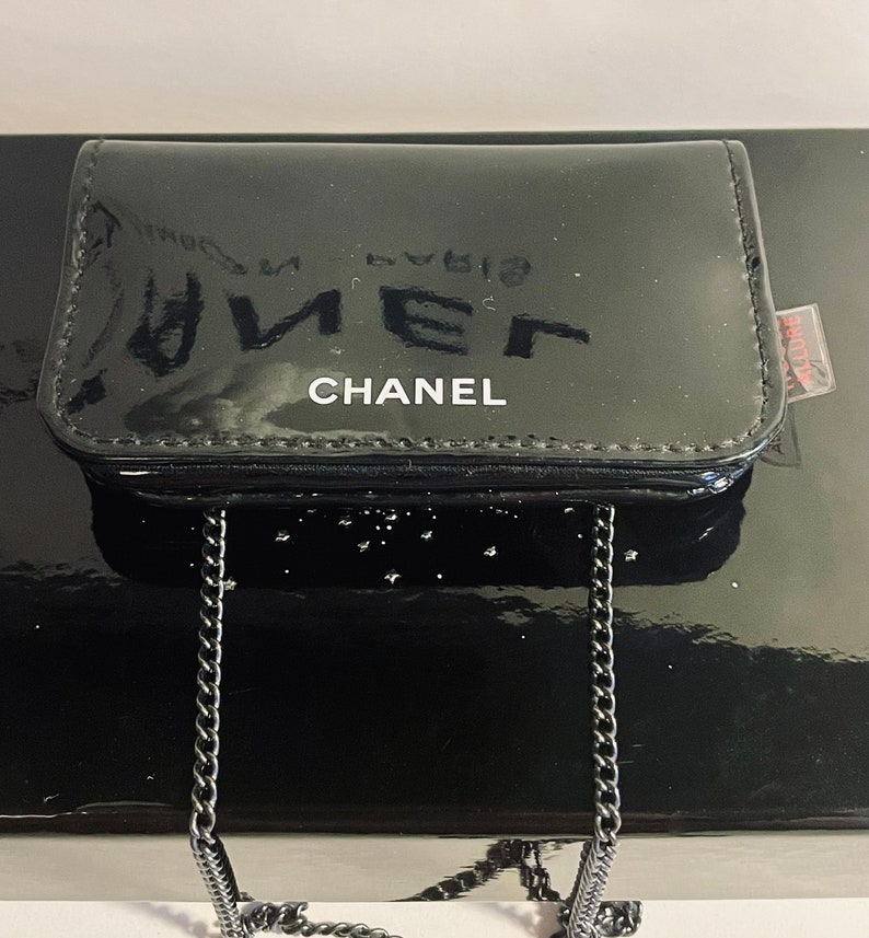 Puede incluir: Un monedero negro brillante con una cadena plateada. La palabra "CHANEL" est&aacute; impresa en blanco en la parte delantera. El monedero es rectangular y est&aacute; sobre una superficie negra reflectante.