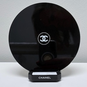 May include: A black, circular compact mirror with a white Chanel logo. The mirror is set on a black, geometric base with the brand name in white lettering. The design is sleek and modern.