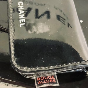 Puede incluir: Un estuche de maquillaje CHANEL negro brillante con las palabras "ROUGE ALLURE" en una peque&ntilde;a etiqueta. El estuche tiene una superficie reflectante, y el logotipo de CHANEL es blanco. La est&eacute;tica general es elegante y lujosa.