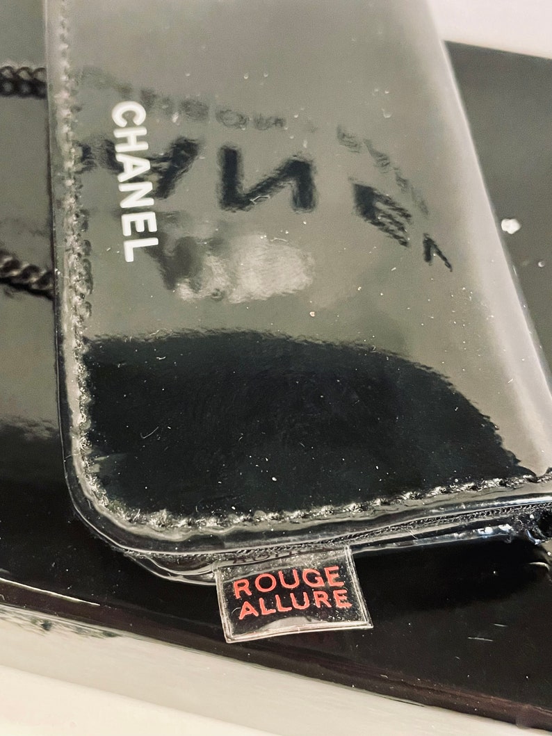 Puede incluir: Un estuche de maquillaje CHANEL negro brillante con las palabras "ROUGE ALLURE" en una peque&ntilde;a etiqueta. El estuche tiene una superficie reflectante, y el logotipo de CHANEL es blanco. La est&eacute;tica general es elegante y lujosa.