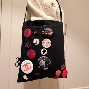 May include: Black tote bag adorned with various round pins in shades of pink, red, and white. The pins feature text and images, including the Chanel logo and the words "rouge coco" and "mademoiselle". The bag has a shoulder strap.
