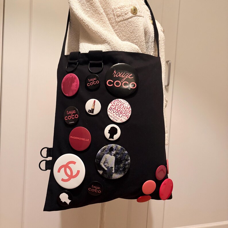 May include: Black tote bag adorned with various round pins in shades of pink, red, and white. The pins feature text and images, including the Chanel logo and the words "rouge coco" and "mademoiselle". The bag has a shoulder strap.