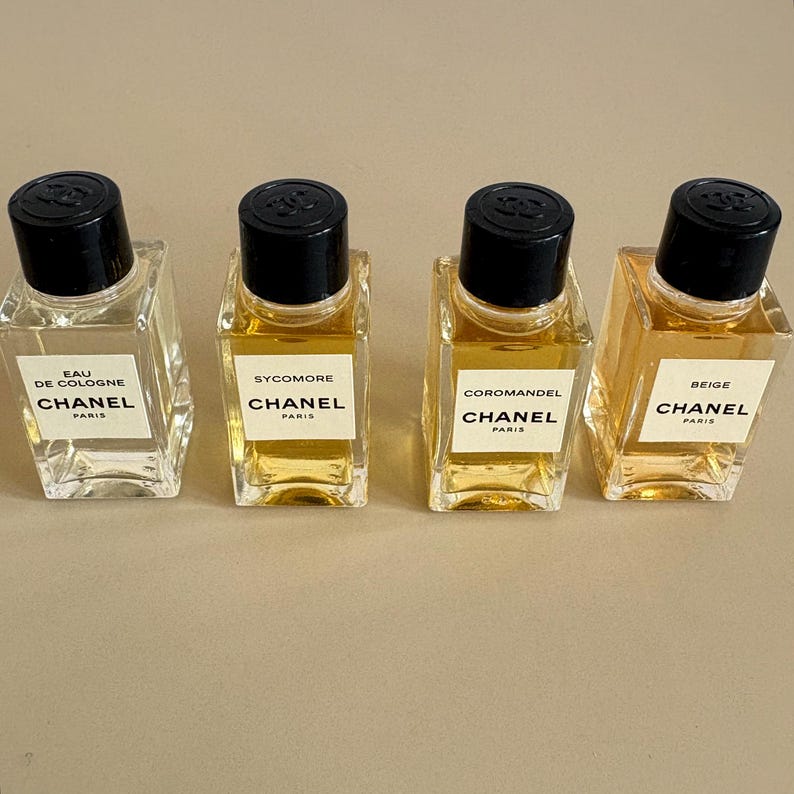 May include: Four Chanel perfume bottles with black caps and white labels. The bottles are filled with different colored perfumes, ranging from clear to amber. The labels read "Eau de Cologne", "Sycomore", "Coromandel", and "Beige".
