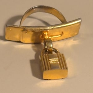 Peut inclure: Une bague en métal doré avec une barre rectangulaire et un petit charme cadenas. La bague a une bande incurvée et une barre rectangulaire avec un petit charme cadenas suspendu. Le cadenas a des lignes verticales et une petite boucle.