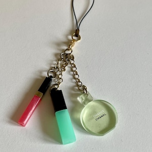 Könnte beinhalten: Ein goldener Ketten-Handy-Anhänger mit drei Anhängern: einem rosa Lippenstift, einem grünen Nagellack und einem runden, durchsichtigen Fläschchen mit dem Text "Chance Chanel".