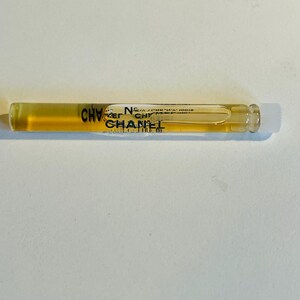 May include: A clear glass vial containing a golden-colored liquid, with the word "CHANEL" printed in black and gold. The vial has a white cap and is filled with a liquid. The vial is on a white surface.