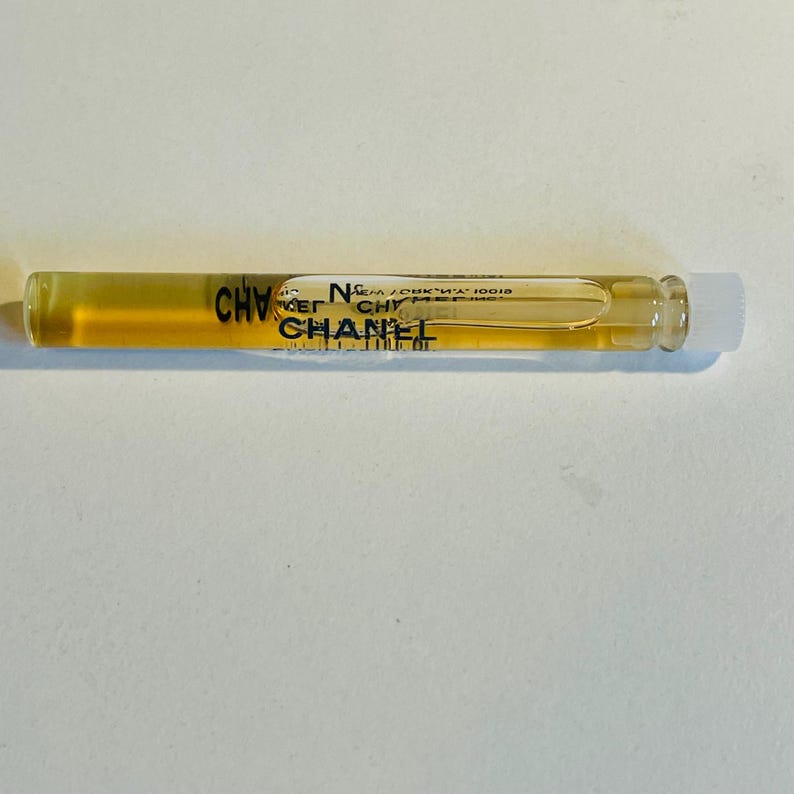 May include: A clear glass vial containing a golden-colored liquid, with the word "CHANEL" printed in black and gold. The vial has a white cap and is filled with a liquid. The vial is on a white surface.