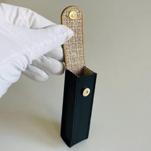 Op de afbeelding: Een donker marineblauwe rechthoekige etui met een gouden drukknoopsluiting. De binnenkant is gevoerd met een geweven beige en gouden stof. De etui wordt vastgehouden door een gehandschoende hand.