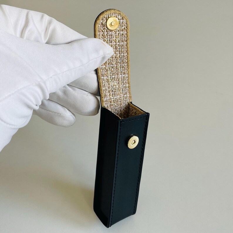 Op de afbeelding: Een donker marineblauwe rechthoekige etui met een gouden drukknoopsluiting. De binnenkant is gevoerd met een geweven beige en gouden stof. De etui wordt vastgehouden door een gehandschoende hand.