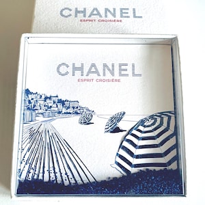 Könnte beinhalten: Eine weiße Schachtel mit dem CHANEL-Logo und dem Text "ESPRIT CROISIERE". Die Schachtel zeigt eine blau-weiße Illustration einer Strandszene mit Sonnenschirmen, einer Küstenlinie und einem Pier.