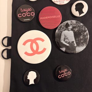 May include: A collection of round Chanel-themed buttons in various designs. Some buttons feature the "rouge coco" text, the Chanel logo, and the word "Mademoiselle". Other buttons have a silhouette of a woman's head or a black and white photo.