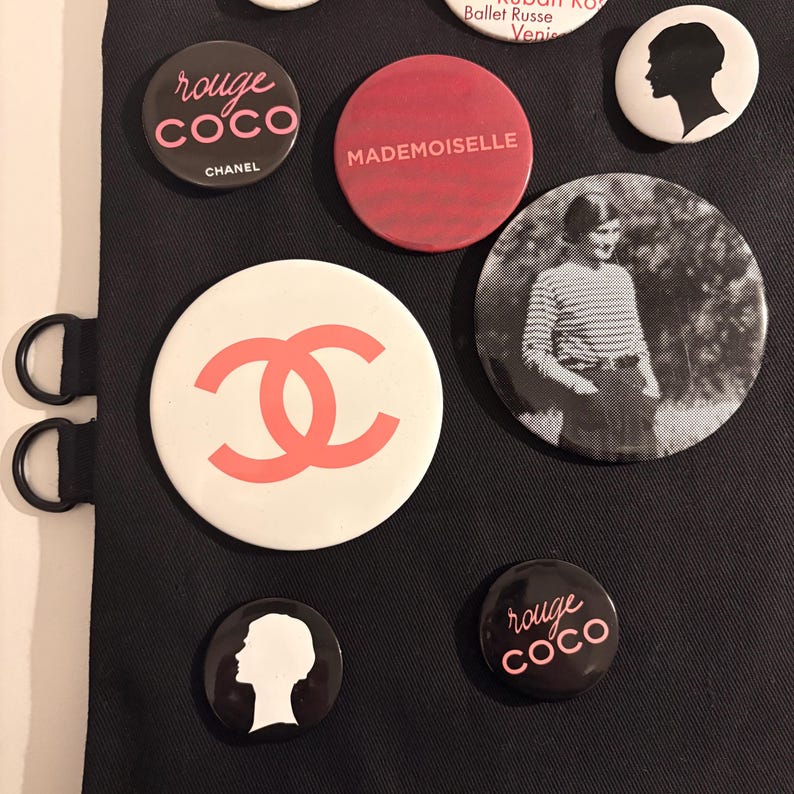 May include: A collection of round Chanel-themed buttons in various designs. Some buttons feature the "rouge coco" text, the Chanel logo, and the word "Mademoiselle". Other buttons have a silhouette of a woman's head or a black and white photo.