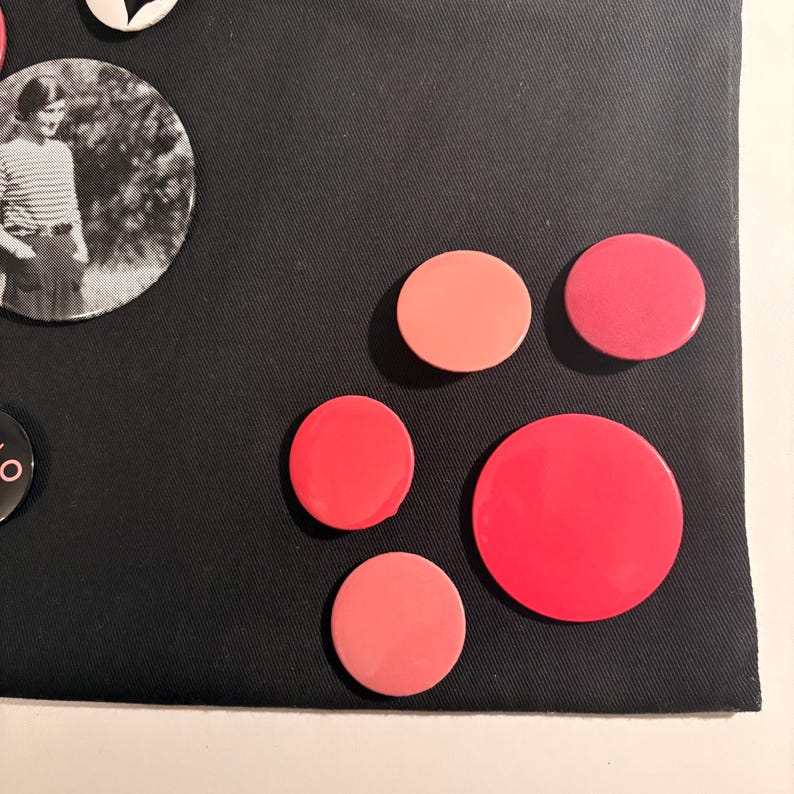 May include: A collection of colorful round buttons in shades of pink and red, arranged on a black fabric surface. One button features a black and white photo of a person. The buttons vary in size and color.
