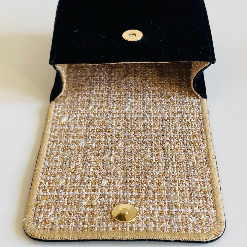 Op de afbeelding: Open zwart fluwelen etui met een goudkleurige druksluiting. De binnenkant heeft een geweven stof in tinten goud, beige en wit. De randen zijn afgewerkt met een gouden rand. De etui is ontworpen om kleine items in te bewaren.