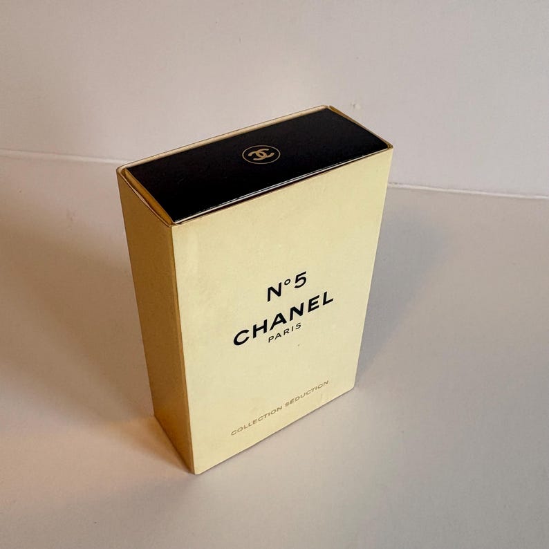 May include: A rectangular perfume box with a black top and a cream-colored body. The box features the text "N&deg;5 CHANEL PARIS" in black, with the Chanel logo on the top. The words "COLLECTION SEDUCTION" are printed at the bottom.