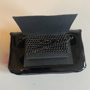 Puede incluir: Un peque&ntilde;o bolso de mano negro brillante con una solapa doblada, que revela un collar de cadena de metal oscuro. El bolso de mano tiene forma rectangular con bordes redondeados. Una peque&ntilde;a etiqueta roja y blanca es visible en el lateral.