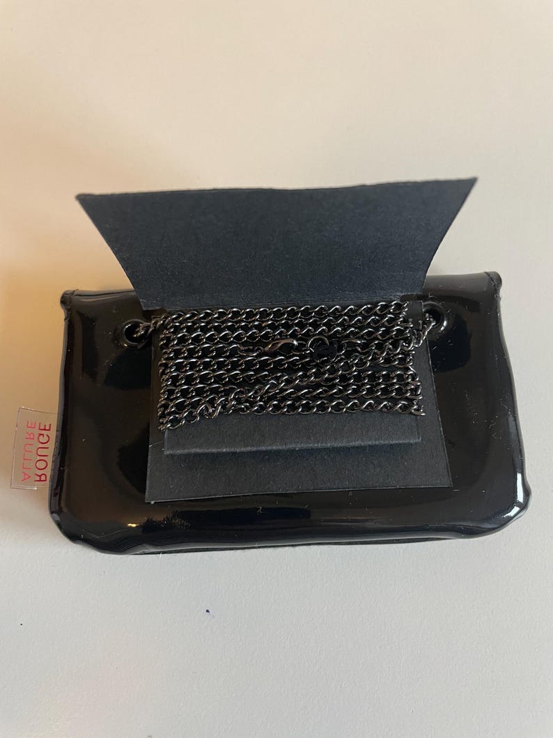 Puede incluir: Un peque&ntilde;o bolso de mano negro brillante con una solapa doblada, que revela un collar de cadena de metal oscuro. El bolso de mano tiene forma rectangular con bordes redondeados. Una peque&ntilde;a etiqueta roja y blanca es visible en el lateral.