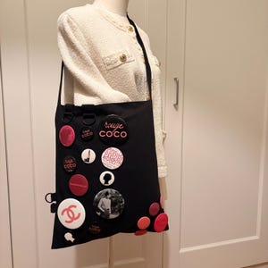 May include: Black tote bag adorned with various round pins in red, white, and black. The pins feature text and graphics, including the words "rouge COCO" and a double "C" logo. The bag has a long black strap.