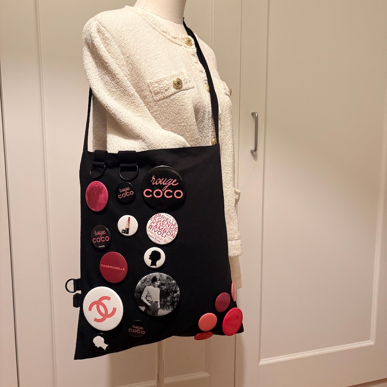 May include: Black tote bag adorned with various round pins in red, white, and black. The pins feature text and graphics, including the words "rouge COCO" and a double "C" logo. The bag has a long black strap.