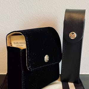 Op de afbeelding: Een zwart fluwelen cosmetica-etui met een zilveren knoopsluiting en een kleine tube Sublimage Le Fluide. Een zwart leren etui met een zilveren knoop staat naast het cosmetica-etui. Beide etuis hebben een zilveren logo.