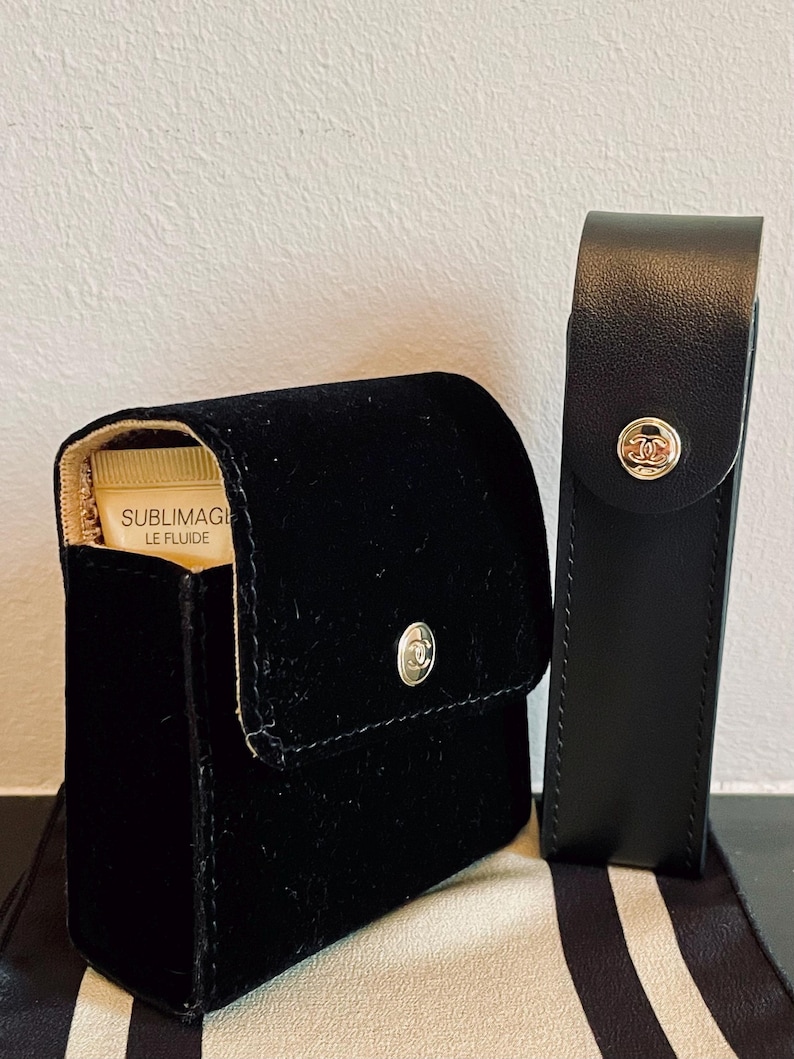 Op de afbeelding: Een zwart fluwelen cosmetica-etui met een zilveren knoopsluiting en een kleine tube Sublimage Le Fluide. Een zwart leren etui met een zilveren knoop staat naast het cosmetica-etui. Beide etuis hebben een zilveren logo.