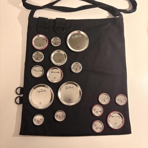 May include: Black tote bag with a shoulder strap, adorned with various sized silver and red-rimmed pinback buttons. The buttons are arranged across the front of the bag, creating a unique pattern. The bag has metal rings and loops.