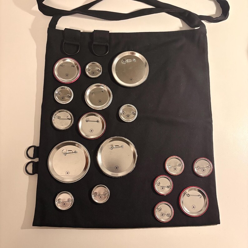 May include: Black tote bag with a shoulder strap, adorned with various sized silver and red-rimmed pinback buttons. The buttons are arranged across the front of the bag, creating a unique pattern. The bag has metal rings and loops.