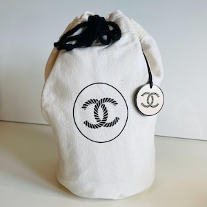Puede incluir: Una bolsa de lona blanca con cordón de ajuste de cuerda negra. La bolsa presenta un gran logotipo negro en un círculo y una etiqueta más pequeña a juego. El logotipo es un diseño de doble C entrelazado.