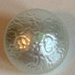 May include: A round, iridescent button with a swirling, textured surface. The button has a light green and gold design in the center, with a light blue and white design on the right side. The button is set against a neutral background.