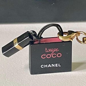 Könnte beinhalten: Ein schwarzer Lippenstift-Anhänger mit einem goldenen Band und einer kleinen schwarzen Box mit den Worten "rouge COCO CHANEL" in Rosa und Weiß. Der Anhänger hat eine schwarze Kordel und einen goldenen Verschluss.