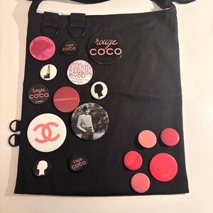 May include: Black tote bag adorned with various round pins. The pins feature the "rouge coco" logo, a silhouette, and a photo of a woman. Other pins are in shades of pink and red.