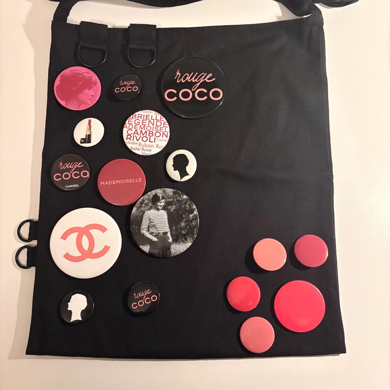 May include: Black tote bag adorned with various round pins. The pins feature the "rouge coco" logo, a silhouette, and a photo of a woman. Other pins are in shades of pink and red.