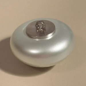 May include: A pearlescent, light-colored button with a metallic shank. The button has a rounded, slightly flattened shape. The shank is a small, circular piece of metal with a loop for sewing. The button is likely used for clothing or crafts.