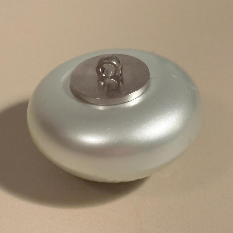 May include: A pearlescent, light-colored button with a metallic shank. The button has a rounded, slightly flattened shape. The shank is a small, circular piece of metal with a loop for sewing. The button is likely used for clothing or crafts.