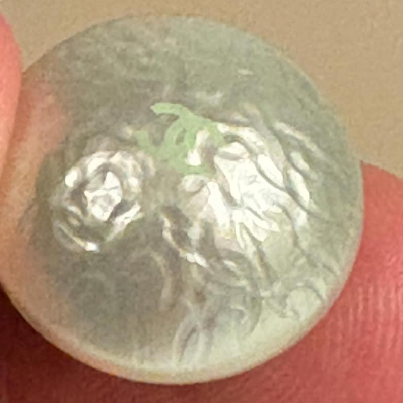 May include: A round, light green button with a floral design. The button has a slightly textured surface and a raised floral pattern in white and green. The button is held between two fingers.