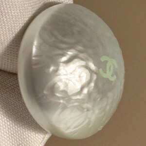 May include: A pearlescent, oval-shaped button with a light green logo on the surface. The button has a swirling, textured design and a subtle sheen. The button is likely for clothing or accessories.