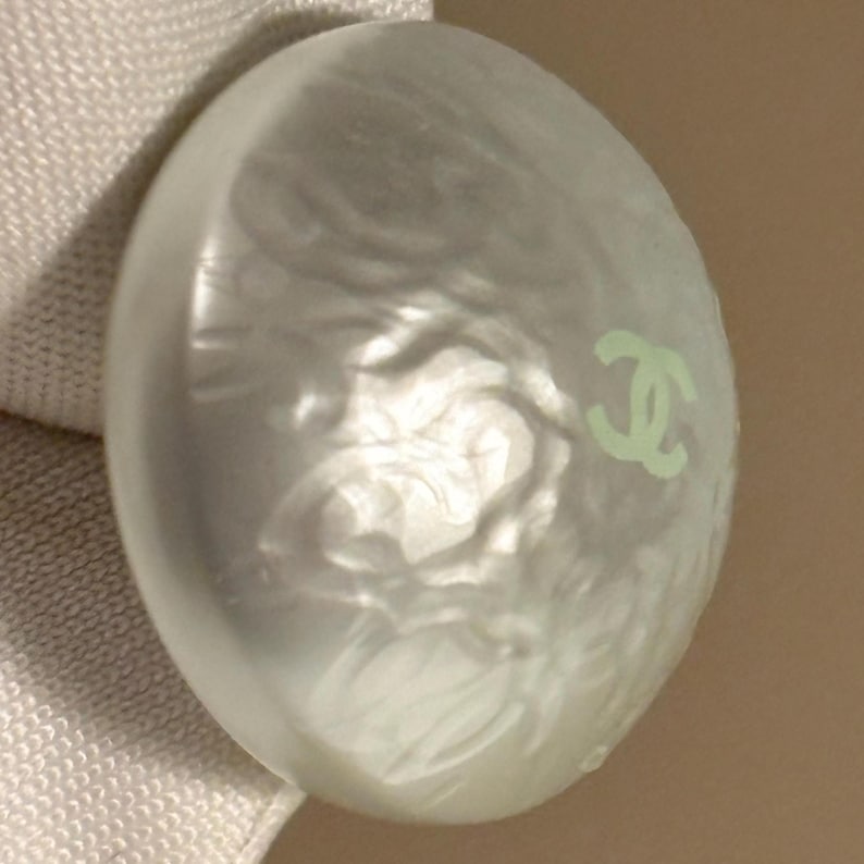 May include: A pearlescent, oval-shaped button with a light green logo on the surface. The button has a swirling, textured design and a subtle sheen. The button is likely for clothing or accessories.