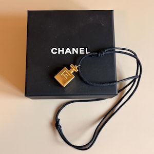 Op de afbeelding: Een goudkleurige hanger in de vorm van een parfumflesje met het Chanel-logo, hangend aan een dun zwart koord. De hanger rust op een vierkante zwarte doos met de witte tekst CHANEL in het midden van het deksel in een schreefloos lettertype.