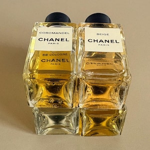 May include: Two Chanel cologne bottles, one labeled "Coromandel" and the other "Beige." Both bottles are rectangular with a clear glass body and a dark blue cap. The labels are white with black text, and the cologne inside is a golden color.