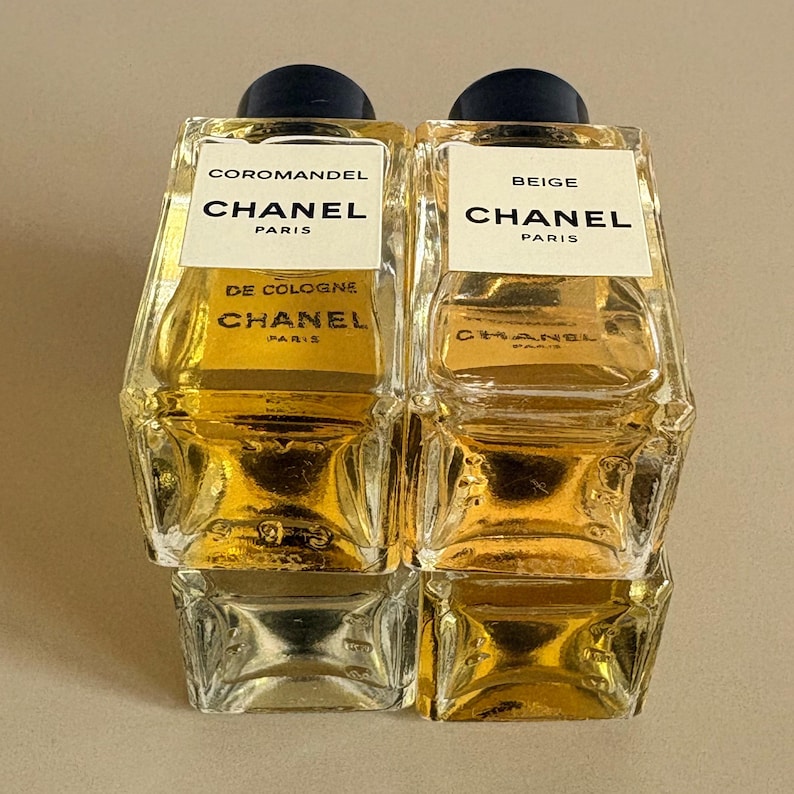 May include: Two Chanel cologne bottles, one labeled "Coromandel" and the other "Beige." Both bottles are rectangular with a clear glass body and a dark blue cap. The labels are white with black text, and the cologne inside is a golden color.